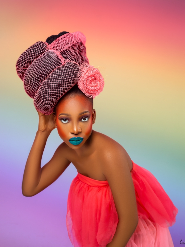 Artistic Portrait Of A Woman With A High Pink Textured Headpiece And Blue Lipstick Against A Rainbow Gradient Background.